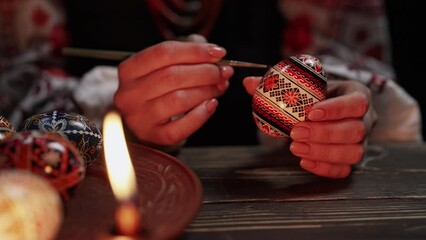 Ukrainian woman painting traditional ornamets on Easter egg - pysanka. Artist working in national costume under candle light. Preparation for Christian holiday