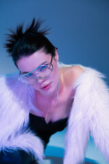 fashionable woman in white faux fur jacket and transparent eyeglasses on blue background with...