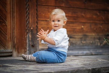 blond one year old baby boy in chic white  shirt looking serious & focused into the camera outside of a rustic wooden hut  while clapping hands and making hand signs