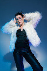 model in black corset and trendy faux fur jacket posing with hands behind head on blue background.