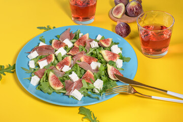 Autumn figs salad with arugula, feta, raw ham in blue plate on yellow background served with ros wine. Flexitarian diet, paleo diet fall salad, close up