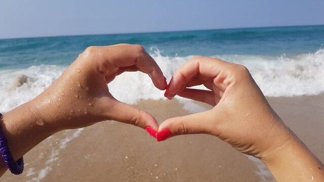 A young woman shows a heart shape with her hands while standing on the shore of the blue Mediterranean sea