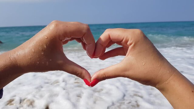 A young woman shows a heart shape with her hands while standing on the shore of the blue Mediterranean sea