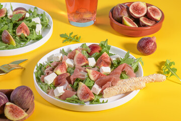 Autumn figs salad with arugula, feta, raw ham in white plate on yellow background served with rose wine and grissini. Flexitarian diet, paleo diet fall salad, close up