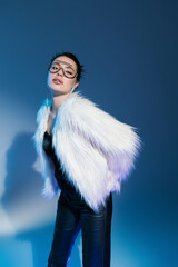 young brunette woman in white faux fur jacket and transparent sunglasses looking at camera while...