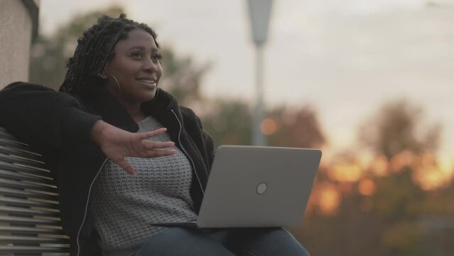 Online meeting. Inspired woman. Video connection. Happy black lady sitting square bench talking on laptop in sunset light.