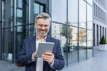 Mature gray-haired investor outside office building using tablet computer, senior businessman in business suit holding tablet reading news smiling.