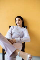 Obraz premium trendy woman in cozy sweater looking at camera while sitting on yellow background.