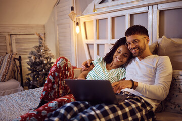 Young happy couple using laptop while relaxing on bed at home during winter holidays.