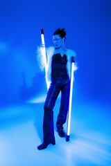 Fototapeta premium Stylish woman in furry jacket and sunglasses holding lamps on blue background.