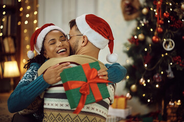 Happy black woman embracing her boyfriend after getting Christmas present from him at home.