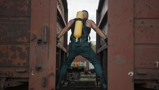 Bodybuilder with balloon drager on his back pulls himself up on his hands between freight wagons. Street workout. Back view