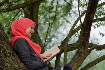 Cute muslim student reading a book while sitting on the tree branch in the park. Modern and fashionable female model representing islam and knowladge, education and stressfree. Arabic culture