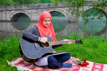 Preaty muslim girl playing acoustic guitar sitting on the river bank with the bridge in the backgroud.