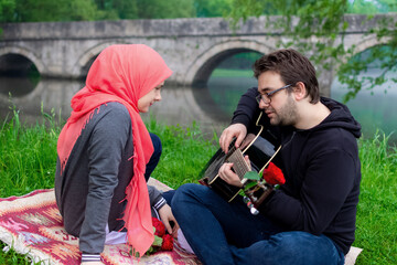 Muslim couple enjoying the day in the nature. Husband playing guitar to his lovely wife while sitting on the river bank with the bridge reflecting in the background