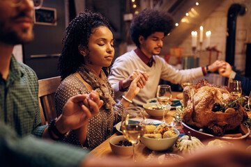 Black woman and her friends praying during Thanksgiving meal at dining table.