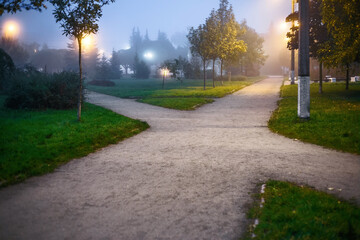 Crossing wide pedestrian sandy alleys in the park among grass and trees at dusk, in the light of night lamps. conceptual landscape