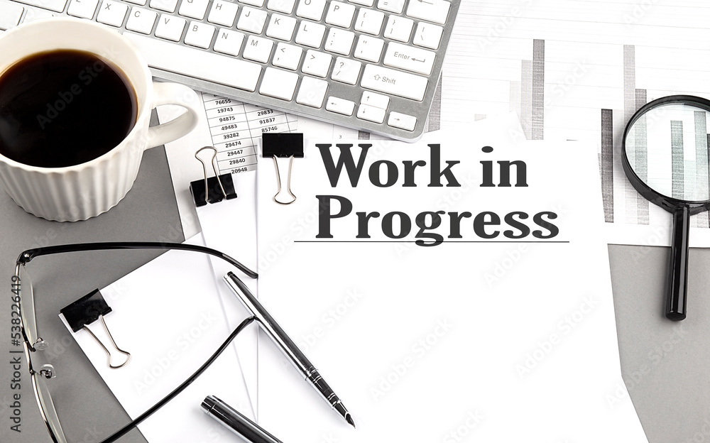 Poster work in progress text on a paper with magnifier, coffee and keyboard on grey background