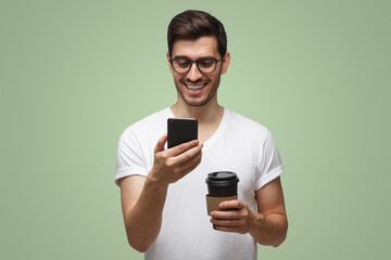 Guy laughing at message holding cellphone and drinking coffee, isolated on green