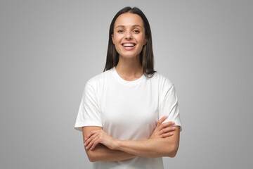 Fototapeta premium Portrait of young smiling female standing in white t-shirt with crossed arms on gray background
