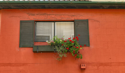 Fototapeta na wymiar Double window with open wooden shutters and flower box against an orange painted wall. The flower box contains a variety of flowers, including bright red geraniums that cascade down the wall. 