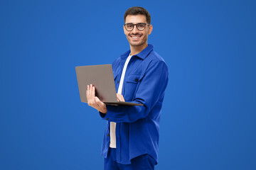 Obraz na płótnie Canvas Young man standing holding laptop and looking at camera with happy smile, isolated on blue
