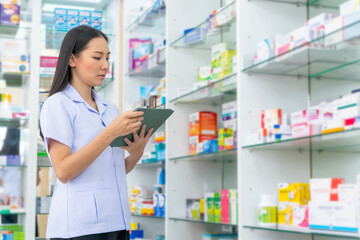 Medical pharmacy and healthcare providers concept. Attractive Asian woman pharmacist working on digital tablet checking stock of medical product, drugs, medicine and supplements on shelf in drugstore.