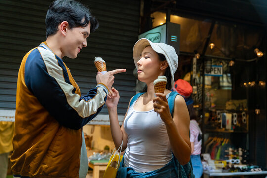 Young Asian couple enjoy and fun outdoor lifestyle shopping at street market on summer holiday vacation. Man and woman couple eating ice cream cone while walking and shopping at weekend street market.