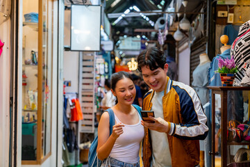 Obraz na płótnie Canvas Asian couple enjoy and fun outdoor lifestyle shopping at street market on summer holiday vacation. Happy man and woman couple using mobile phone together while walking and shopping at weekend market.