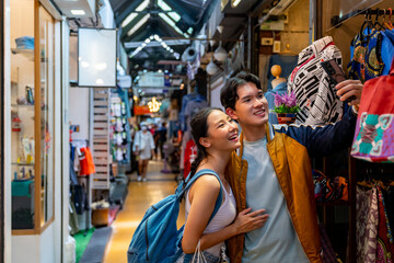 Obraz na płótnie Canvas Asian couple enjoy and fun outdoor lifestyle shopping at street market on summer holiday vacation. Man and woman couple using mobile phone taking selfie together while shopping at weekend market.