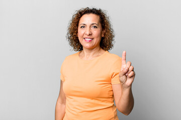 middle age hispanic woman smiling and looking friendly, showing number one or first with hand...