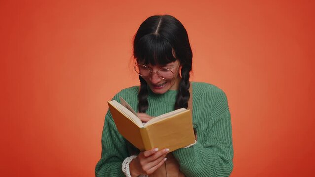 Young nerd woman with pigtails in glasses reading funny interesting fairytale story book, leisure hobby, knowledge wisdom, education, learning, study, wow. Latin girl isolated on orange background