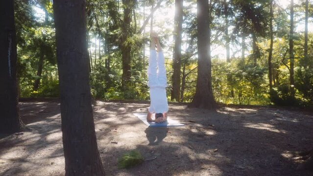 Caucasian adult man dressed in white holds headstand position on a  yoga mat in a pine tree forest with great light. Camera moving around, wide shot.