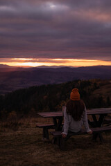 Young woman from behind is sitting at bench and look at sunset mountains view