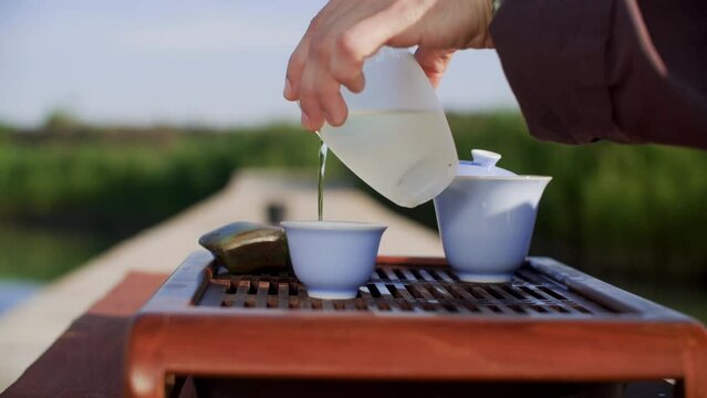 Close-up of cropped man pouring water on tea leaves in cup. Ceramic Gaiwan bowls on chaban tray in sunlight. He is preparing traditional Chinese tea beverage during summer. Oolong tea