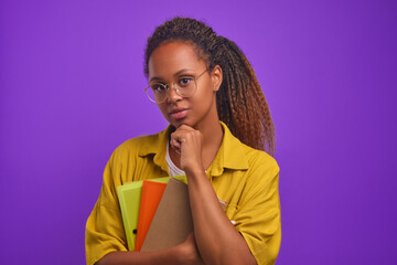 Young serious beautiful African American woman teacher touches chin and holds workbooks with books...