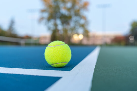 Photo Taken In Late Evening Under Lights Of A Yellow Tennis Ball On Blue Tennis Court With White Line And Green Out Of Bounds.