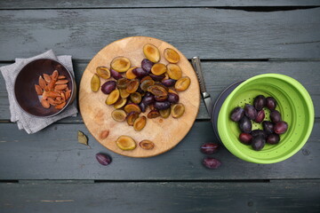 Plums halves on the cutting board with pits in the bowl at side and the colander with whole fruits arranged on the wooden background