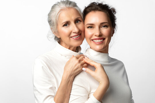 Cosmetic portrait of two women of different ages and generations smiling and holding hands on a white background. A young beautiful woman and her mother looking happily into the frame together