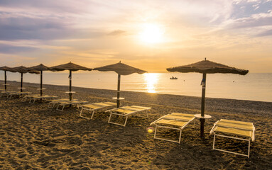  empty beach during beautiful sunrise or sunset with chaise loungues and nice umbrellas with blue sea, sun glow and amazing cloudy sky on thr background