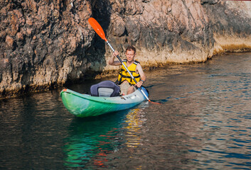 A man swims in a kayak in the summer on the sea against the background of rocks