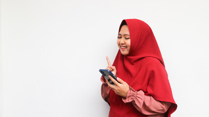 Lovely happy smiling woman wearing red hijab looking and pointing on smartphone over isolated white background