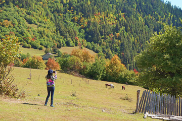 Female hiker taking photos of a herd of horses grazing at the mountain farm, Mestia town, Svaneti region, Georgia