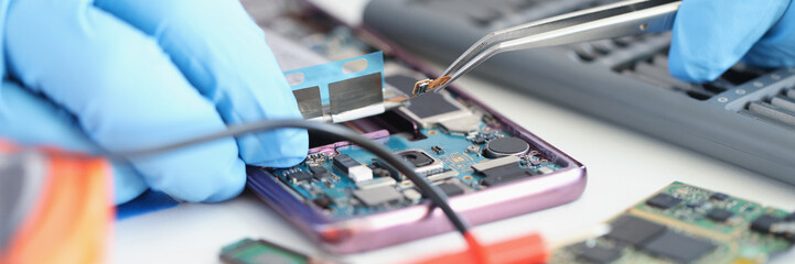 Hands in gloves hold a smartphone microchip with tweezers