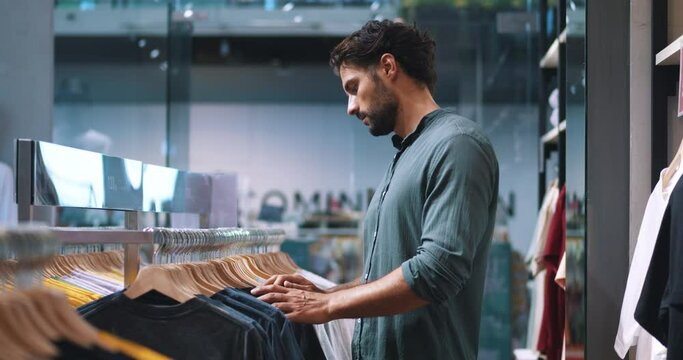 Attractive bearded multi-ethnic macho man in dark-green casual shirt, shopping for t-shirts in fashionable men's clothing store, inspecting differents shirts, price tags and the quality of fabric