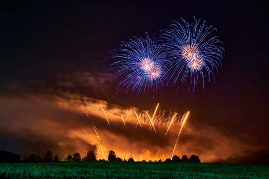 Fireworks with blue stars and yellow fountains against dark night sky. Green meadow and tree line. Ideal for Sylvester and New Year's Day. Strong smoke in the humid air. Germany, Ostfildern.