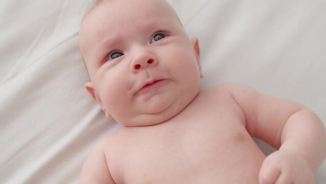 Beautiful newborn baby. Cute two-month-old baby with bare belly looks into camera, slow motion. Active newborn boy lies on the bed and moves his arms. High angle view of a chubby male baby..