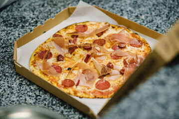 Pizza in a cardboard box on table. Top view of hot tasty pepperoni pizza sprinkled grated cheese topping. Food home delivery. Baked traditional Italian pizza with meat, prosciutto, salami. Self-pickup