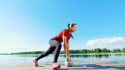 Beautiful, athletic young blond woman doing different exercises with weights, dumbbells, lunges, squats. Lake, river, blue sky and forest in the background, summer sunny day. High quality photo