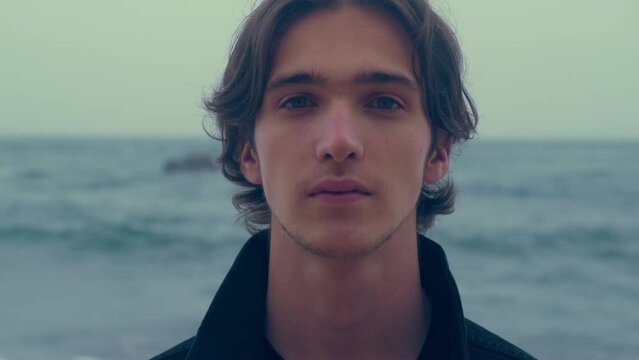 Young man  on the empty ocean beach, on the cloudy rainy weather. Confident and calm guy is on a sandy shore looks at the camera. Closeup face of handsome man posing outdoors. Slow motion 4k footage.